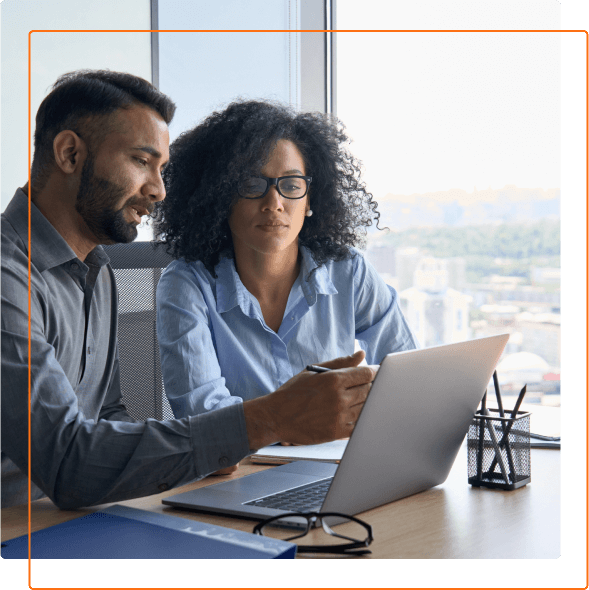 Two professionals discuss ideas in a modern office setting, working collaboratively on a laptop with a city view in the background.