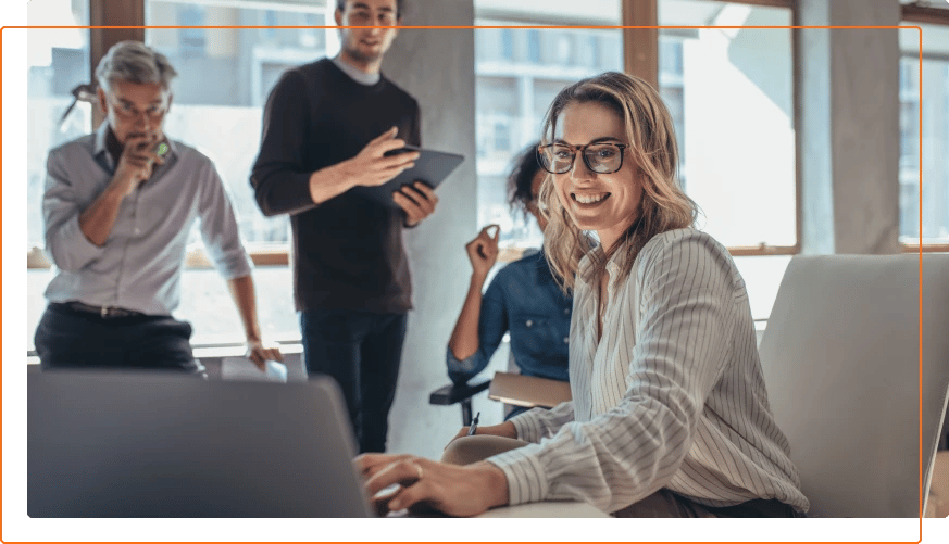 Photo of a woman smiling while working at her desk with colleagues watching.