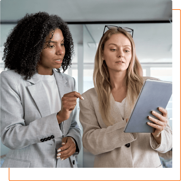 Two women in business attire are discussing and reviewing information on a tablet in a modern office setting.