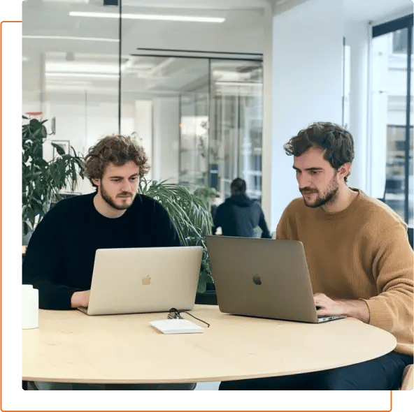 Two people sit at a round table working on laptops in a modern office space, surrounded by plants and natural light.