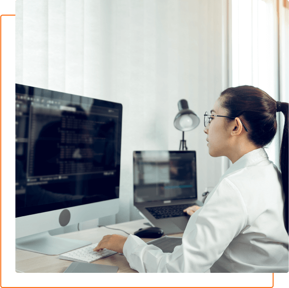 A person in a white lab coat works on computers in a bright, modern workspace, focusing on digital data on the screens.