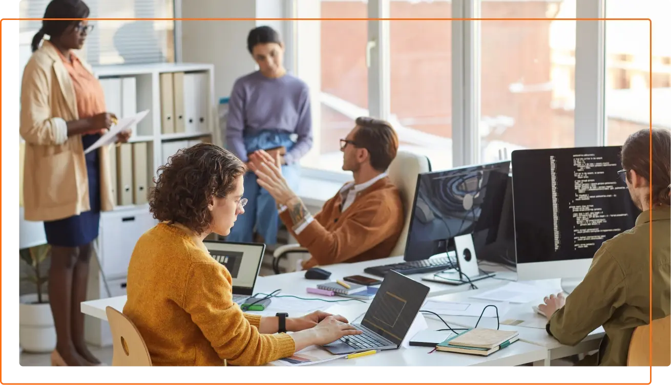 A diverse group of professionals engages in a collaborative meeting in a bright office, with computers and notes on the table.