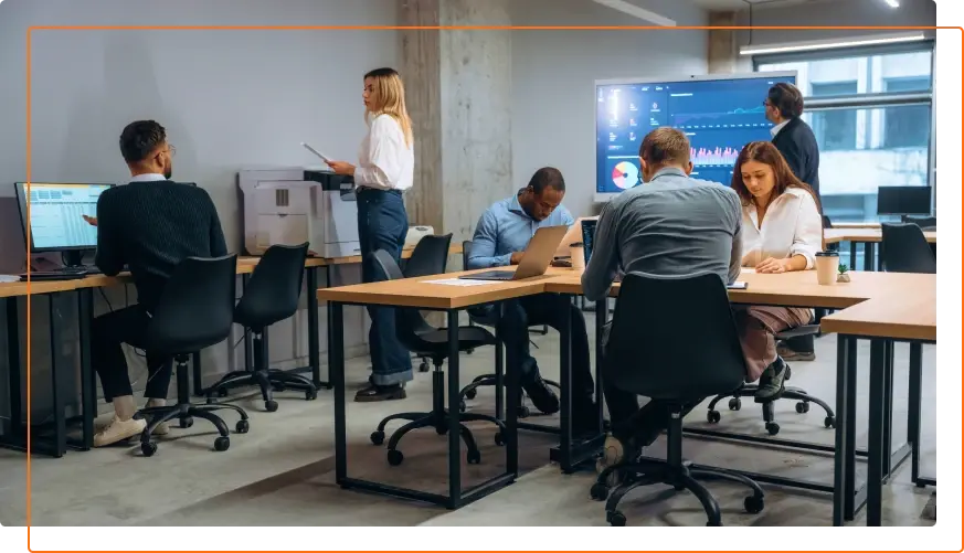 A modern office environment with several individuals collaborating at desks, using computers and engaging in discussions.