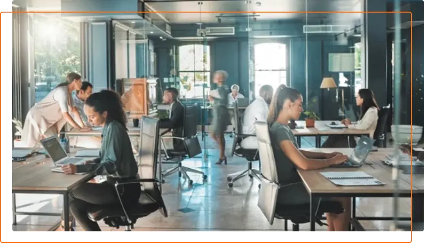A modern office environment with people working at desks, using laptops, and engaging in discussions, illuminated by natural light.