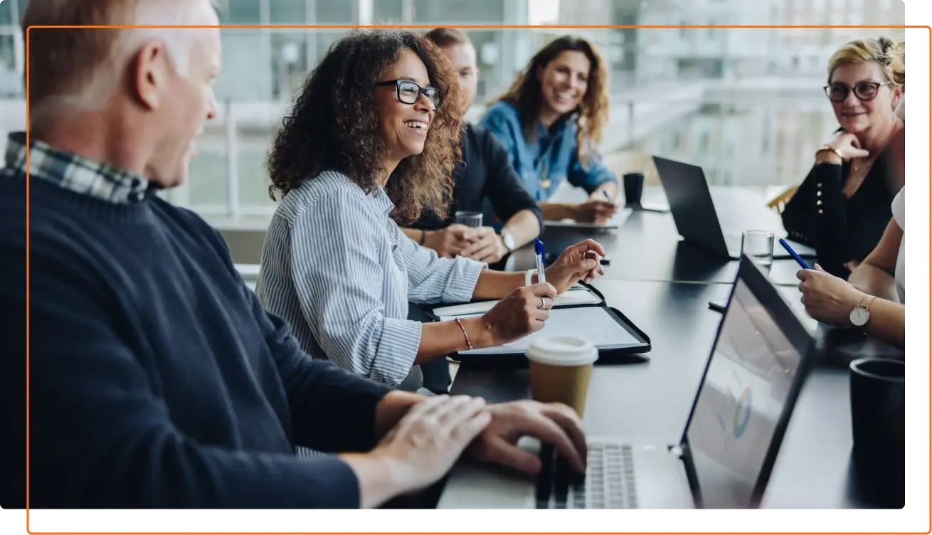 A diverse group of people engaged in a collaborative meeting around a conference table, with laptops and coffee cups present.